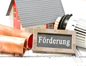 Ein Haus mit einem Schild, wo Förderung drauf steht, daneben ein Thermostat und Kupferheizrohre