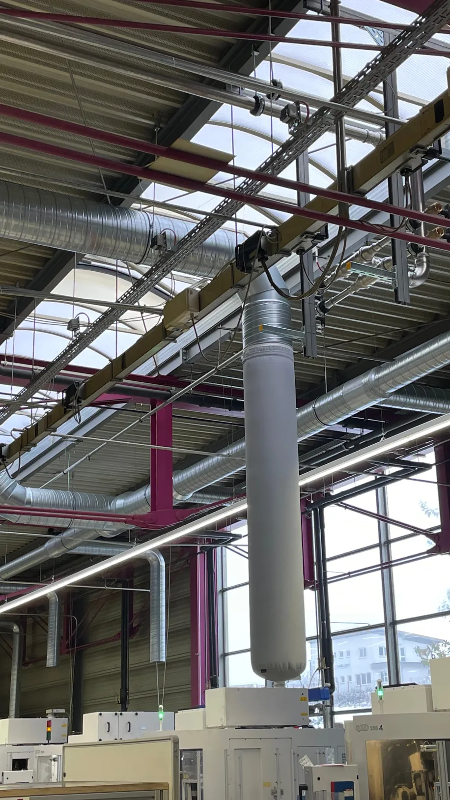 In einer modernen Produktionshalle sind textile Luftauslässe an einem komplexen System aus Lüftungsrohren und Kabeltrassen unter der Hallendecke installiert.