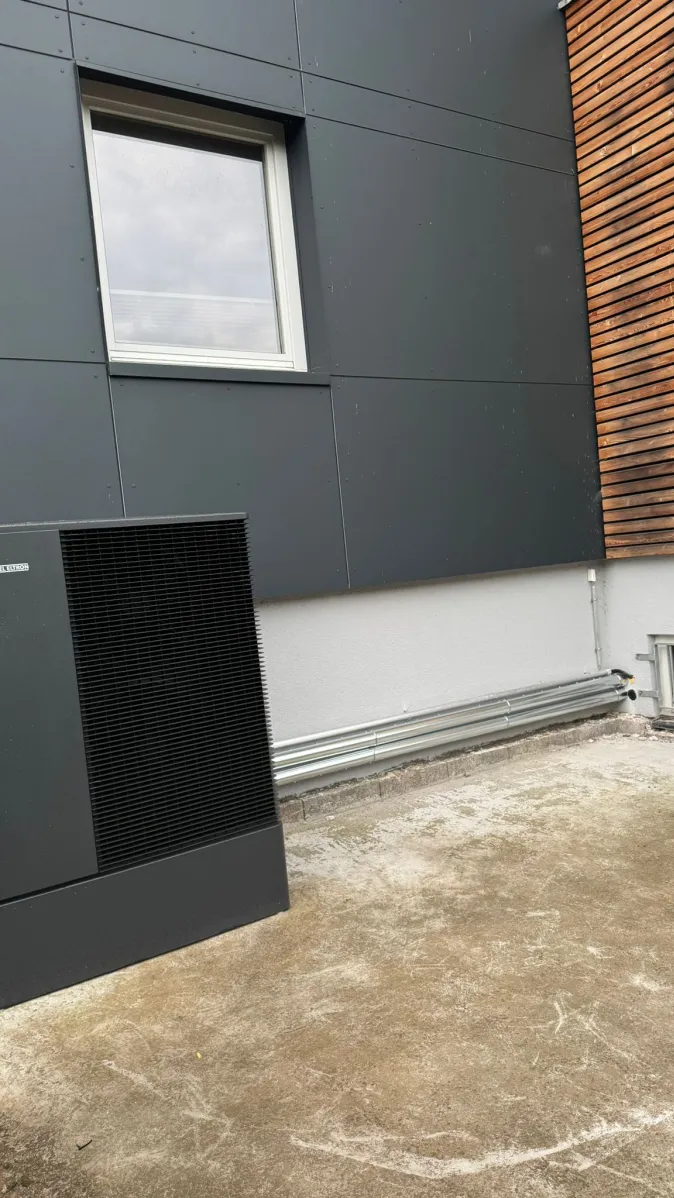 Eine moderne Luft-Wasser-Wärmepumpe in dunklem Gehäuse steht neben einem Wohnhaus mit dunkler Fassadenverkleidung und Holzpaneelen.