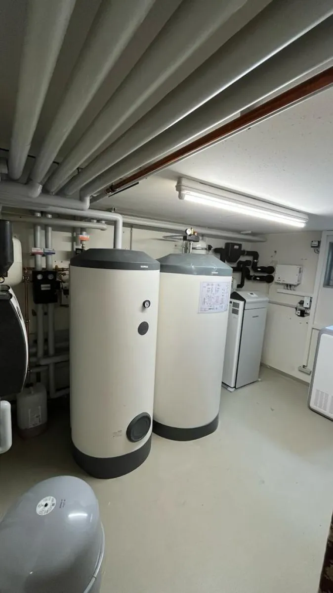 Ein moderner Heizungsraum zeigt eine komplette Wärmepumpen-Innenanlage mit zwei großen, weißen Pufferspeichern und umfangreicher Rohrverrohrung an der Decke.