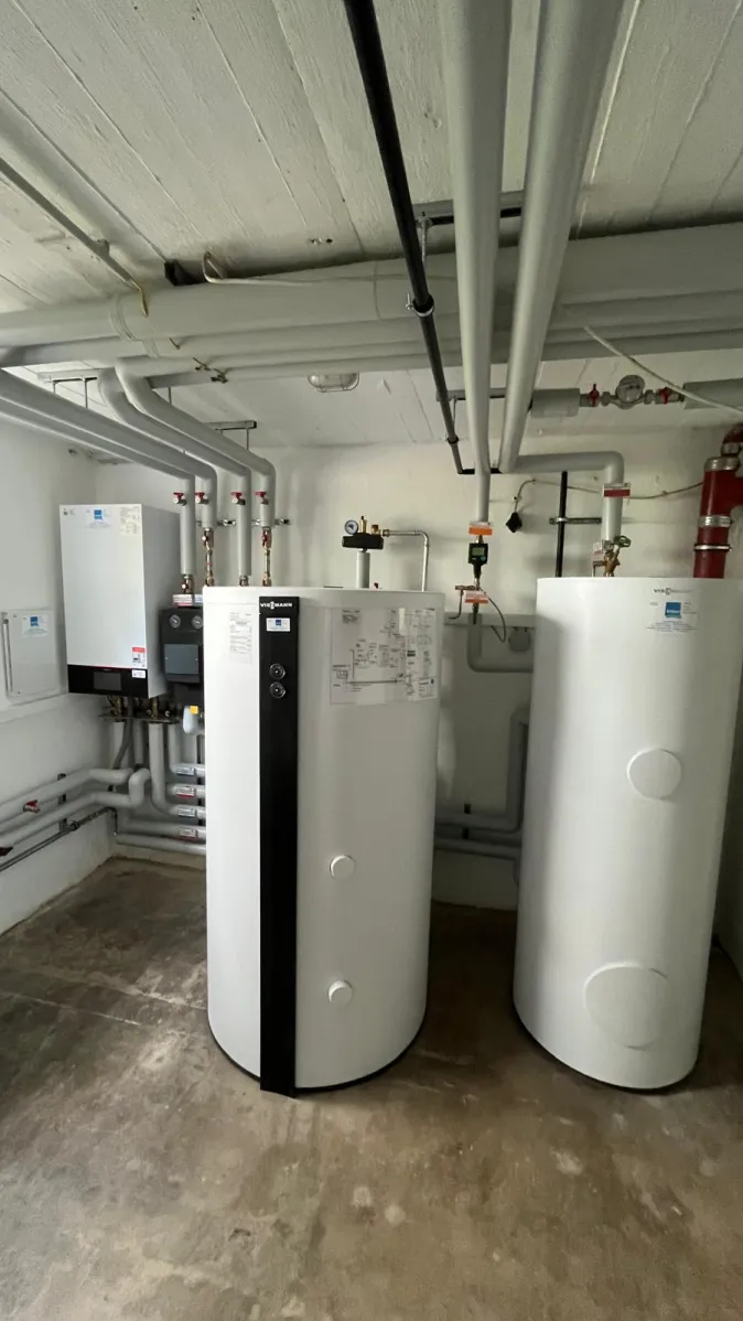 Ein sauber installierter Heizungsraum zeigt zwei große, weiße Pufferspeicher, mehrere Wandgeräte und zahlreiche isolierte Rohrleitungen an Decke und Wänden.