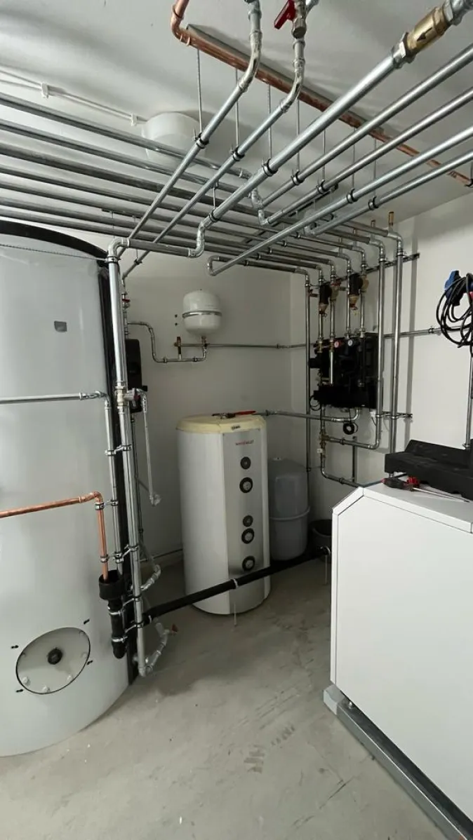 In einem Technikraum sind mehrere Heizkomponenten installiert, darunter ein großer Pufferspeicher, ein Warmwasserspeicher von Weisshaupt und eine moderne Wärmepumpeneinheit.