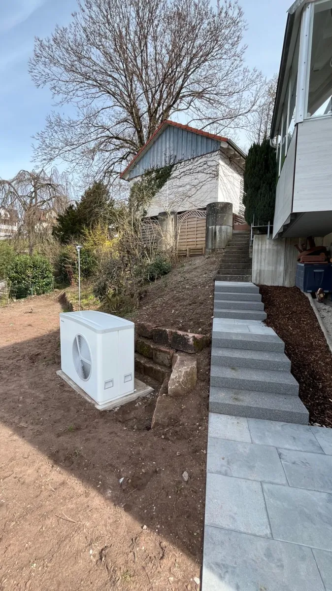 Eine neu installierte Wärmepumpen-Außeneinheit steht auf einem Erdgrundstück neben einem modernen Einfamilienhaus.
