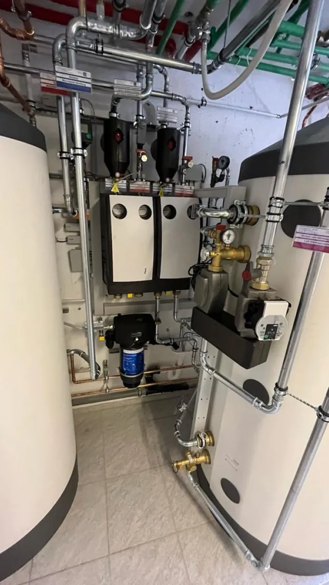 Zwei große weiße Pufferspeicher stehen in einem Technikraum und sind über ein komplexes Rohrsystem mit einer zentralen Pumpen- und Steuerungseinheit verbunden.