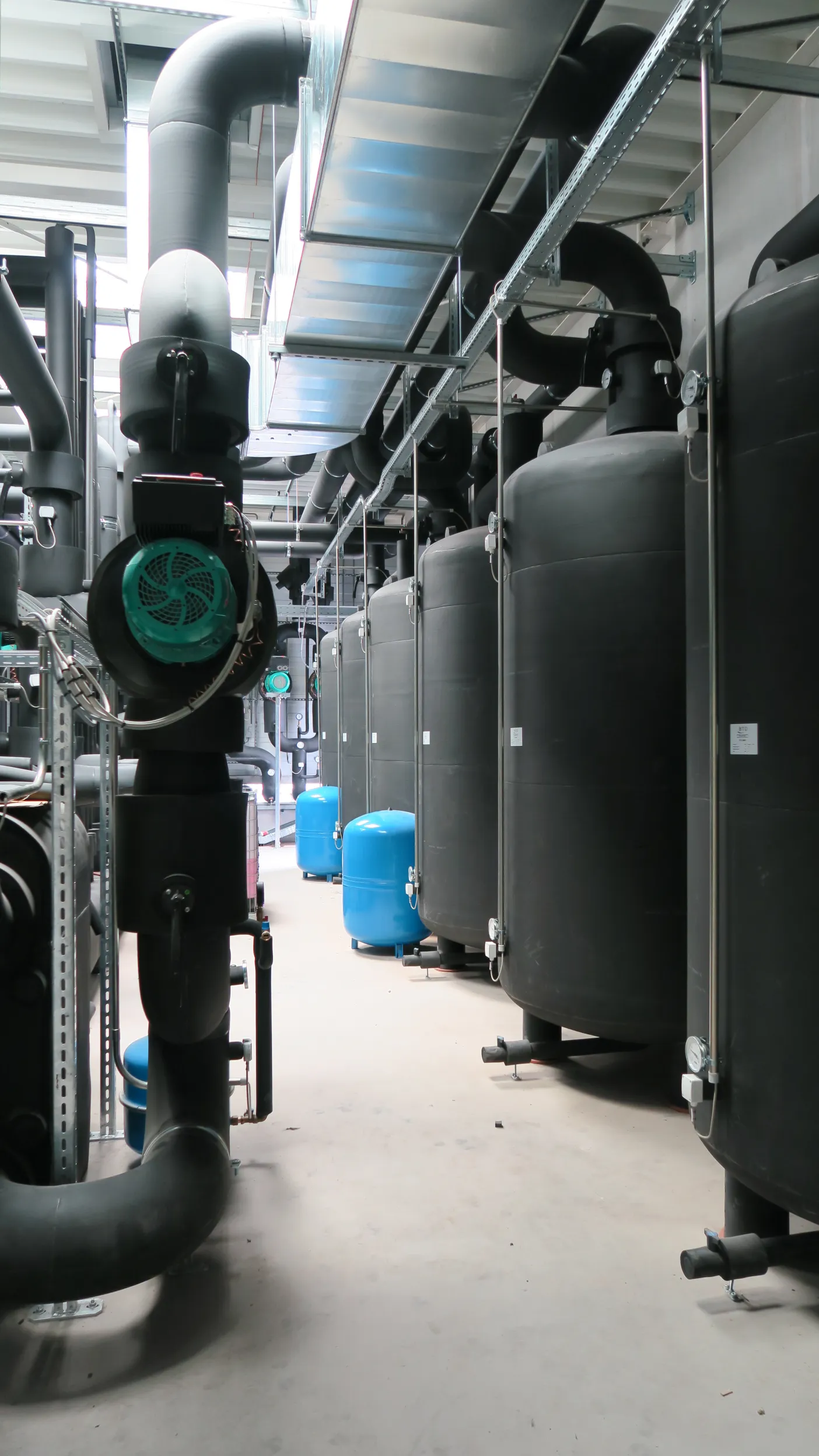 Die Aufnahme zeigt eine Reihe großer schwarzer Pufferspeicher in einem modernen Technikraum, umgeben von komplett isolierten Rohrleitungen.