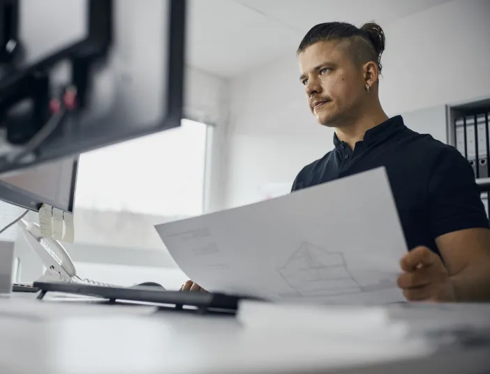 Ein junger technischer Systemplaner sitzt an einem Schreibtisch und hält einen Bauplan in den Händen.