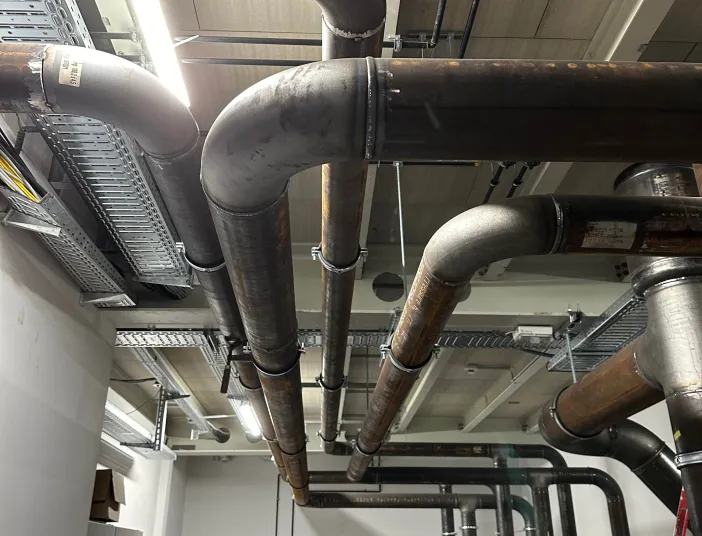 Große Industrierohrleitungen einer Kälteanlage verlaufen an der Decke eines Technikraums, mit Befestigungen, Verteilerleitungen und sichtbarer Installation.