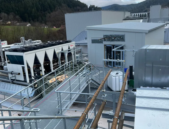 Dachinstallation einer Industrieanlage mit mehreren großen Kältemaschinen, Rohrleitungen und Wartungsstegen vor einer Berglandschaft.