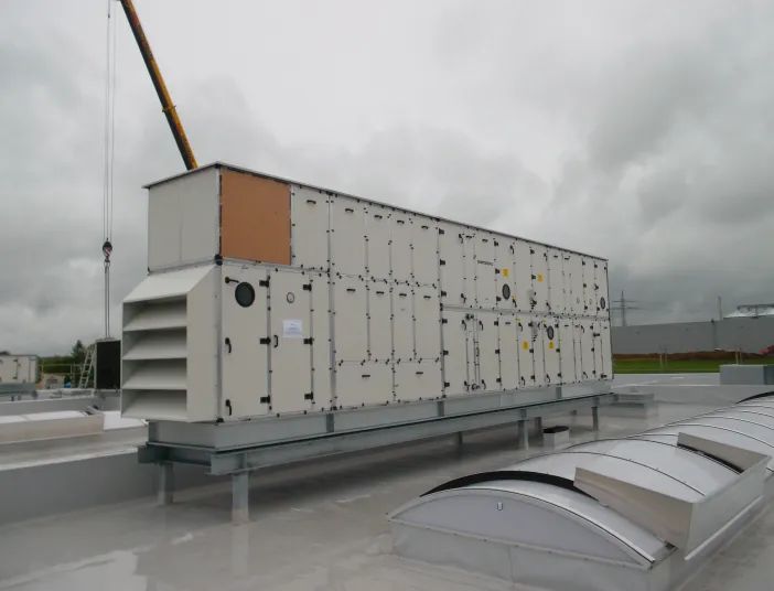 Großes Lüftungs- und Klimagerät auf einem Flachdach, montiert mit Kranunterstützung, vor bewölktem Himmel.