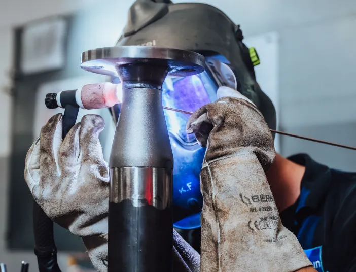 Handwerker mit Schutzhandschuhen und Schweißhelm beim präzisen Schweißen eines Metallrohrs.