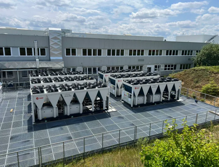 Außenansicht einer Industrieanlage mit mehreren großen Kälte- und Klimageräten der Marke Trane, aufgestellt auf einem Gitterboden vor einem modernen Gebäudekomplex unter blauem Himmel.