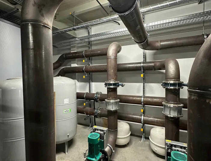 Technikraum einer Kälteanlage mit großen Rohrleitungen, grünen Umwälzpumpen und Druckausdehnungsgefäßen.