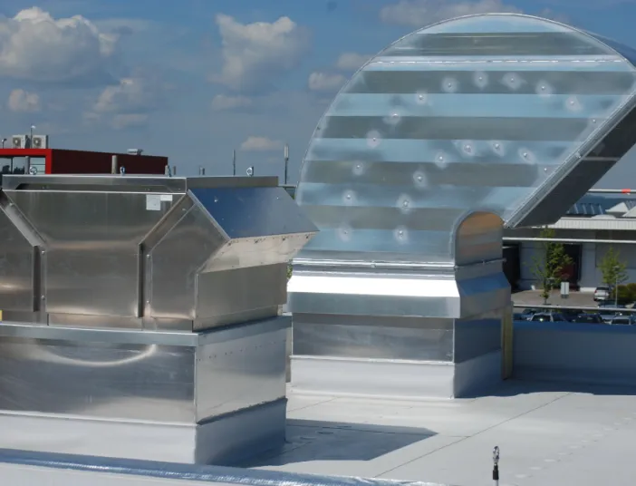 Dachansicht mit großen Lüftungs- und Klimaanlagen aus Metall: Luftkanäle und Abluftsysteme in glänzender Ausführung, im Hintergrund Industriegebäude und blauer Himmel.