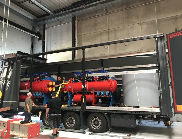 Arbeiter verladen große rote Industriekomponenten auf einem Lkw innerhalb einer Werkshalle. Die Anlage ist mit blauen Armaturen und gelben Anschlüssen versehen und wird mit Spanngurten gesichert.