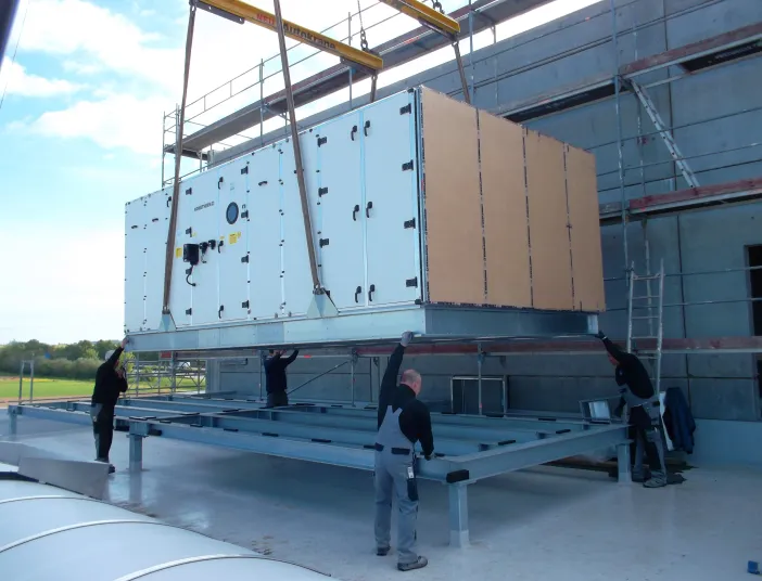 Montage einer großen Lüftungs- bzw. Klimaanlage mit Kran auf einem Gebäudedach, unterstützt von mehreren Monteuren.