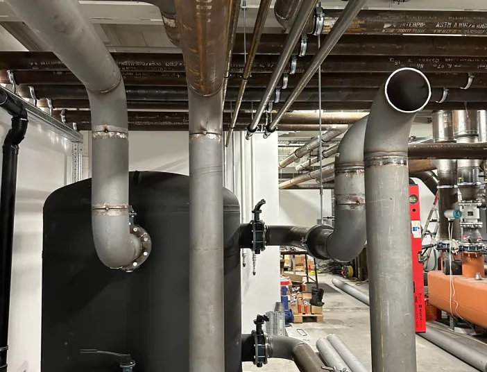 Industrielle Rohrleitungen und schwarzer Pufferspeicher einer Kälteanlage in einem Technikraum, mit sichtbaren Ventilen und Befestigungen an der Decke.