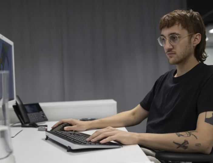 Ein Student mit Brille und Tattoos sitzt konzentriert an einem Schreibtisch vor zwei Monitoren in einem modernen Arbeitsraum.
