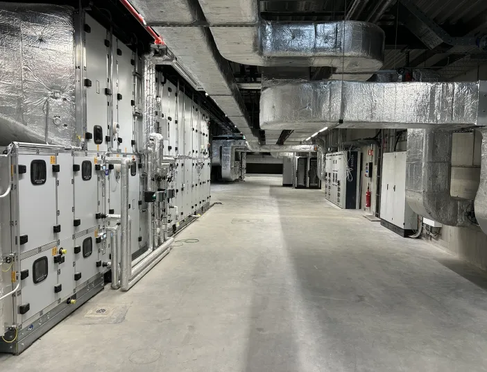 Ein moderner Maschinenraum mit großflächiger Lüftungsanlage zeigt zahlreiche silberne Luftkanäle und technische Anlagen entlang der Wand.