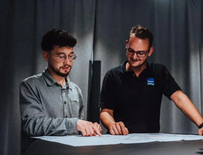 Ein Heizungsverkaeufer der Firma Braun im schwarzen Poloshirt erklärt gemeinsam mit einem Kollegen technische Details auf einem Bauplan.