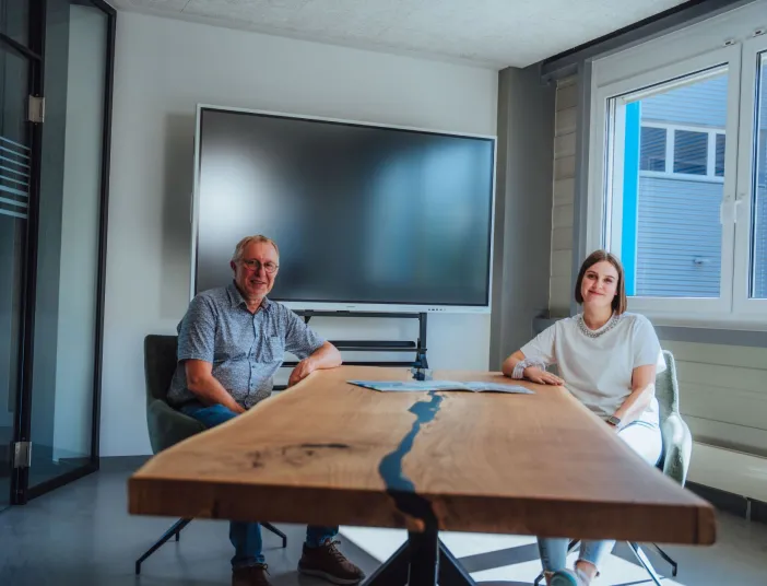 Eine Frau und ein Mann sitzen laechelnd in einem Beratungsraum an einen großen Holztisch.